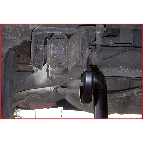 Nahaufnahme der Unterseite eines Autos. Dunkle Metallteile, ein schwarzes Werkzeug und verschiedene Rohre sind zu sehen.